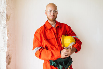 Wall Mural - Young foreman in orange work clothes and protective eyewear holding hardhat and tools in hand happily looking in camera over white background