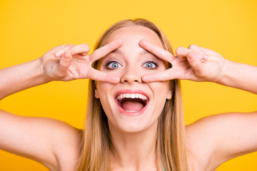 Close up portrait of happy and young woman on yellow background with blond hair, toothy beaming smile and wide open eyes. Happy girl showing v-sign with her two hands and looking at camera