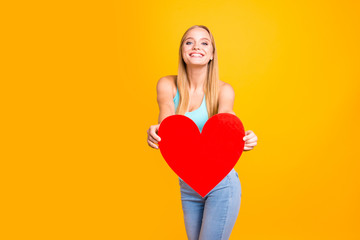 Wall Mural - Happy and cute young blond woman expresses her feelings holding red heart in her hands isolated on yellow background