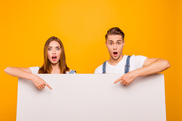 Close up portrait of funny playful lovely young cute couple pointing with fingers to white blank board, showing surprising over yellow background