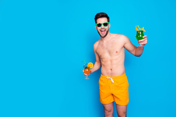 Beach style! Attractive and cheerful man in yellow shorts and eyeglasses, holds cocktails with small umbrella in hands, isolated on blue background. Concept of joyful hot summer