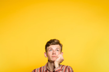 Wall Mural - bored disinterested weariful indifferent unenthusiastic man looking up. hand under his chin. portrait of a young guy on yellow background pop up or peek out from bottom. copy space for advertisement.