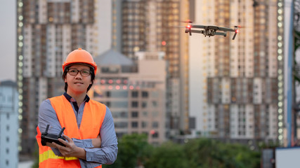 Wall Mural - Young Asian engineer man flying drone over construction site during sunset. Using unmanned aerial vehicle (UAV) for land and building site survey in civil engineering project.