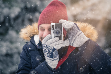 Fashionable man taking pictures with retro camera on snowy day. Winter vacation travel concept.