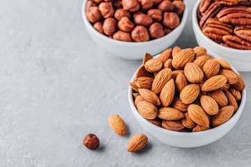 Canvas Print - Almonds, pecans and hazelnuts in white bowls on grey background. Assortment of nuts.