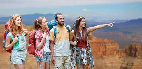 Wall Mural - travel, tourism, hike and adventure concept - group of smiling friends with backpacks pointing finger to something over grand canyon national park background