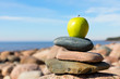 Stones stacked in pyramid, with green apple on top. On large stone on sea shore.Selective focus