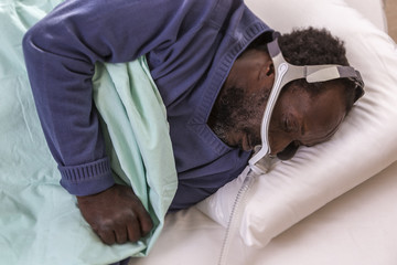 Exhausted afro-american man with sleep apnea using CPAP machine, wearing headgear mask connecting to air tube