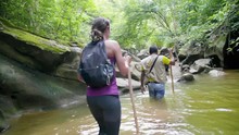 Woman Wearing Athletic Clothing Following A Man Wading Through A River Or Stream In A Rainforest Jungle.
