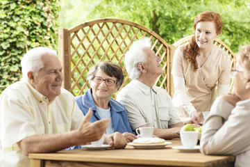 Wall Mural - Group of elderly pensioners enjoying their time together by a table outside in a garden of a retirement home. Young caretaker assisting.