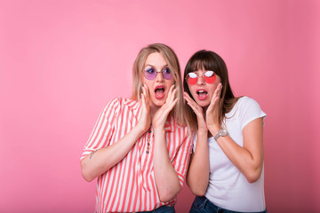 Two cheerful excited young women shouting over pink background.Studio shot of two scared Caucasian female student. raising brows and keeping hands on faces