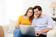 © W PRODUCTION - cheerful young couple on sofa at home websurfing and shopping online with laptop computer internet technology