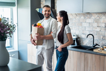 Wall Mural - cheerful couple with paper package full of fresh vegetables standing in kitchen at home