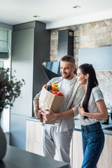 Wall Mural - happy married couple with paper package full of fresh vegetables for dinner in kitchen at home