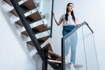 Wall Mural - low angle view of smiling woman in jeans looking away while standing on stairs at home