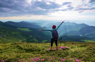 Landscape with the high mountains, the sun rays are coming through the clouds. Forest road. The girl with the back sack stays on the lawn with rhododendrons. Location the Carpathian Mountains.