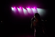 © nagaets - Girl in long gown performing on stage. Girl singing on the stage in front of the lights. Silhouette of singer standing on stage at microphone.