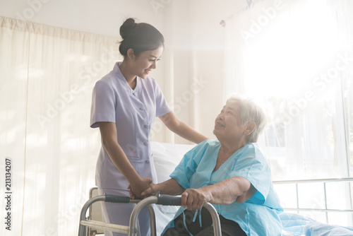 Fotografie, Obraz Asian young nurse supporting elderly patient disabled woman in using walker in hospital