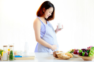 Charming pregnant girl looks her belly and admires her baby in belly. A beautiful woman holds glass of milk and stands at kitchen in her house. Attractive girl feels so happiness and has good health