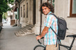© iuricazac - Rear view of young smiling male with curly hair with backpack, looking to the camera posing with bycicle outdoors in the city street. Student riding to college. People, lifestyle, business concept.