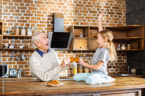 Fotografie, Obraz Joyful aged man eating home made pastry with his granddaughter