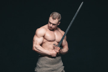 Wall Mural - Bodybuilder man posing with a sword isolated on black background. Serious shirtless man demonstrating his mascular body