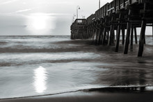 A Black And White Photograph Of The Sandbridge Fishing Pier In Sandbridge Virginia, Virginia Beach During A Sunrise.  A Long Shutter Speed Makes The Water Silky And Dreamy.