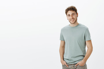 Wall Mural - Portrait of satisfied carefreee attractive male model in casual outfit, holding hands in pockets and smiling with charming expression, tilting head, being happy and in good mood over gray wall