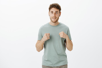 Poster - Portrait of energetic charming european male coworker in trendy t-shirt, raising clenched fists and smiling broadly, ready to give punch or wanting fight, standing against gray background