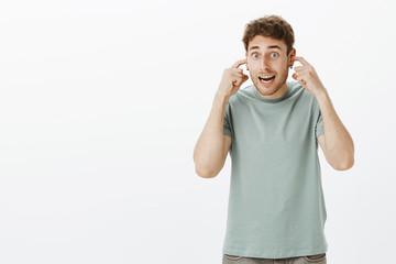 Wall Mural - Guy preparing for big bang, covering ears not hear noise. Excited funny european male student in casual t-shirt, sticking index fingers in ear and waiting for blow while standing over gray wall