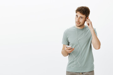 Wall Mural - Handsome confident Caucasian man with bristle, putting on earphones and holding smartphone, picking song and getting ready to go for walk with music in ears, standing self-assured over grey wall