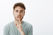© Cookie Studio - Studio shot of pissed off angry young male student in t-shirt and earrings, saying shh and showing shush gesture with forefinger over mouth, demanding keep secret or stay silent over grey wall