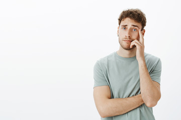 Wall Mural - Unimpressed careless attractive young male with short fair hair, holding hand on cheek and staring with no care at camera, hearing stupid ideas during interview, standing uninterested over grey wall