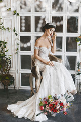 Beautiful and fashionable brunette model girl in stylish wedding dress with naked shoulders and with diadem on her head sits on the chair with bouquet of flowers and posing at luxury vintage interior