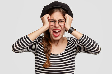 Wall Mural - Depressed beautiful young woman dressed in French style, keeps hands on head, looks desperately at camera, feels worried and stressed, has many problems, stands against white studio blank wall.