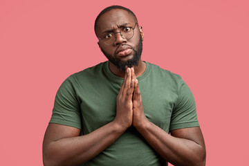 Wall Mural - Photo of serious African American male with pleading expression, keeps palms pressed together, asks for something his lover, hopes to recieve positive answer dressed in casual outfit, isolated on pink