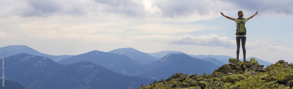 Back view of young slim backpacker tourist girl with raised arms standing on rocky top against bright blue morning sky enjoying foggy mountain range panorama. Tourism, traveling and climbing concept. - obrazy, fototapety, plakaty 
