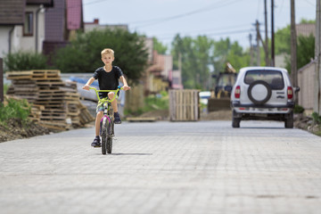 Cute young handsome blond boy in casual summer clothing riding child bicycle along sunny empty paved street on suburb cottages and car blurred background. Children games, fun and safety concept.