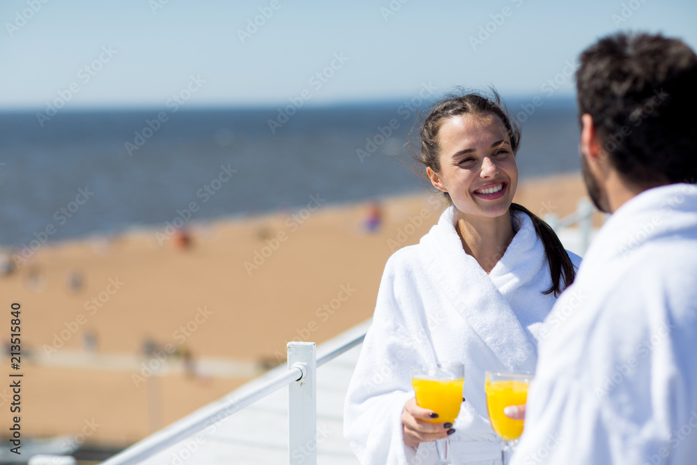 Cherful young wife with glass of juice looking at husband while talking to him at spa resort on sunny day - obrazy, fototapety, plakaty 