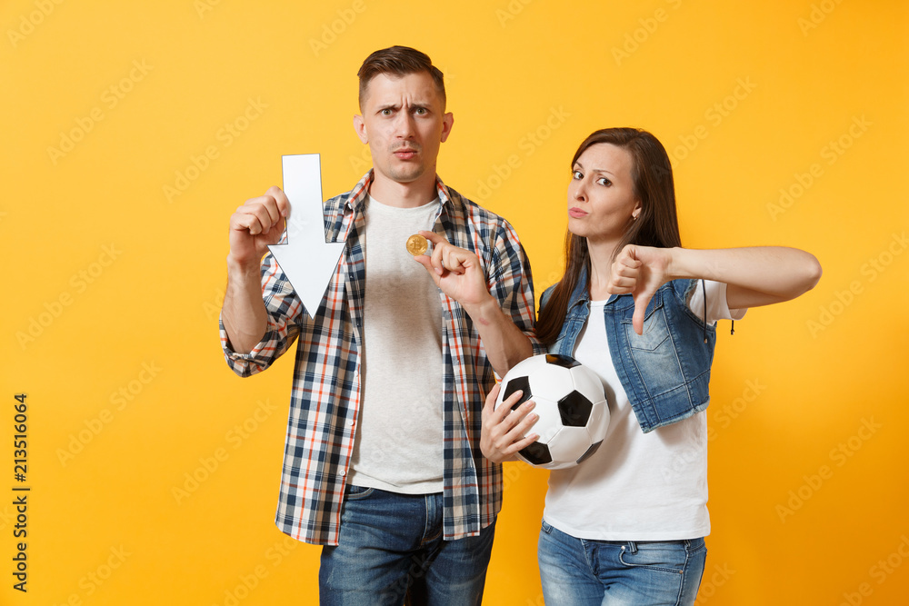 Couple, upset sad woman man, football fans holding bitcoin, metal golden coin, down value fall arrow, soccer ball, cheer up support team isolated on yellow background. Sport family lifestyle concept.