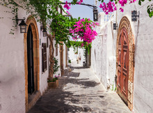 Narrow Street In Lindos Town On Rhodes Island, Dodecanese, Greece. Beautiful Scenic Old Ancient White Houses With Flowers. Famous Tourist Destination In South Europe