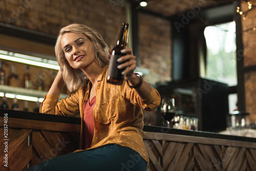 Have a drink. Pleasant blonde woman sitting at the bar counter and raising her beer bottle as if inviting to drink together with her