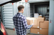 © pressmaster - Rear view of young man in checkered shirt carrying packaged box from car to open container where storing things after home selling