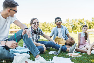 Wall Mural - happy multiethnic teenagers sitting and laughing while studying together in park