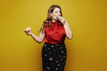 Plus size sexy model, fashionable blonde girl with bright makeup, in red satin blouse and black skirt, with donuts in her arm posing with closed eyes in studio at yellow background