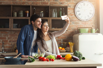 Wall Mural - Happy couple cooking and taking selfie in kitchen