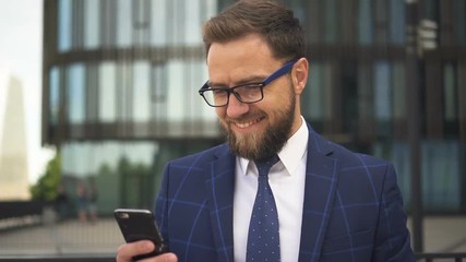 Handsome businessman with beard and in glasses smiling looking to the screen of his smartphone in business district. Outdoors. Portrait.