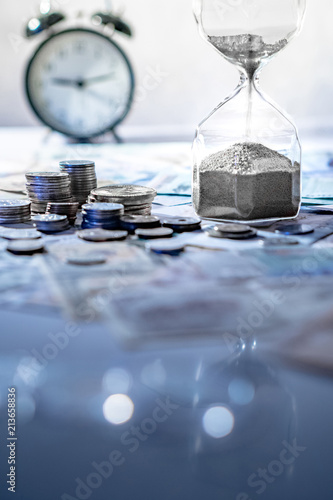 Cuadros en Lienzo Sand running through the shape of hourglass on table with banknotes and coins of international currency