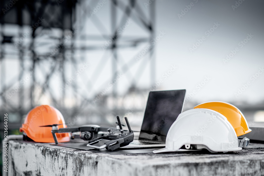 Drone, remote control, smartphone, laptop computer and protective helmet at construction site. Using unmanned aerial vehicle (UAV) for land and building site survey in civil engineering project. - obrazy, fototapety, plakaty 