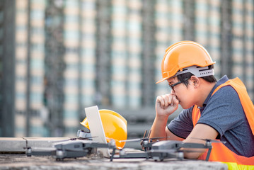 Wall Mural - Young Asian engineer man working with drone laptop and smartphone at construction site. Using unmanned aerial vehicle (UAV) for land and building site survey in civil engineering project.